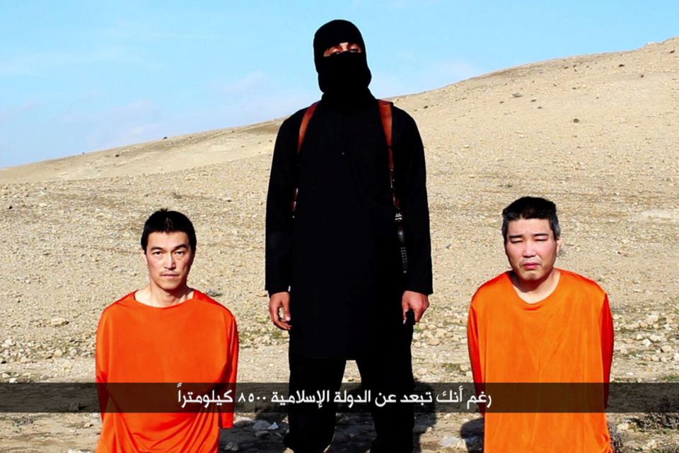 A file image grab taken off a video uploaded on January 20, 2015, reportedly released by the Islamic State (IS) group through Al-Furqan Media, one of the Jihadist platforms used by the militant organisation on the web, allegedly shows Japanese hostages Kenji Goto (L) and Haruna Yukawa (R) in orange jumpsuits with a black-clad militant brandishing a knife as he addresses the camera in English, standing between them at an undisclosed location.