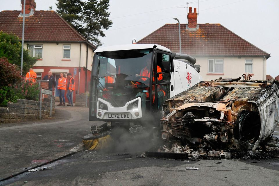 Council workers clear debris around a car that was set alight in Ely, Cardiff, following the riot that broke out after two teenagers died in a crash. Photo: PA Wire