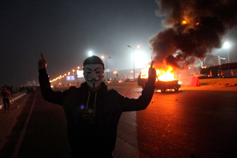 An Egyptian man wearing a mask of the anonymous movement gestures near a burning car outside a sports stadium in a Cairo's northeast district, on February 8, 2015 during clashes between supporters of Zamalek football club and security forces