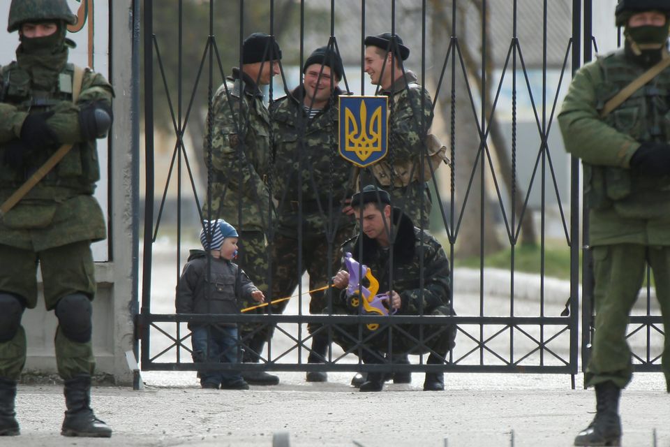 A Ukrainian serviceman plays with a child as men believed to be Russian servicemen stand in front of the gates of a Ukrainian military unit in the village of Perevalnoye outside Simferopol, March 4, 2014. President Vladimir Putin has ordered troops that took part in military exercises in central and western Russia to return to base after completing their training, Russian news agencies quoted the Kremlin spokesman as saying on Tuesday.  REUTERS/David Mdzinarishvili (UKRAINE - Tags: POLITICS MILITARY)